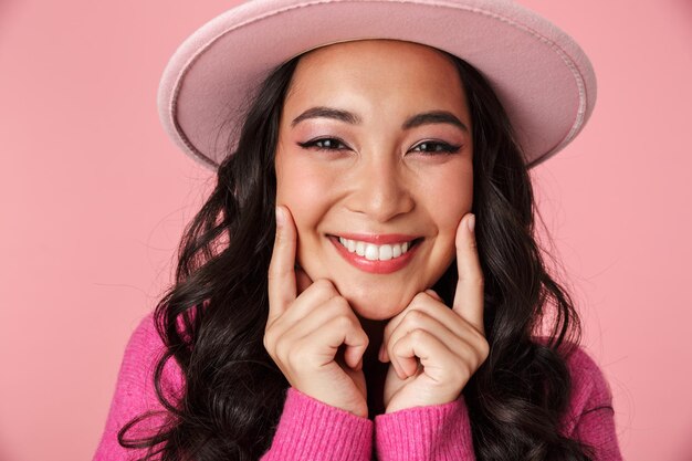 Portret van mode mooi Aziatisch meisje met hoed die haar glimlach toont met vingers geïsoleerd op roze