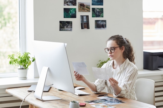 Portret van moderne jonge vrouw met foto's herzien voor publicatie tijdens het werken op pc in wit kantoor, kopie ruimte