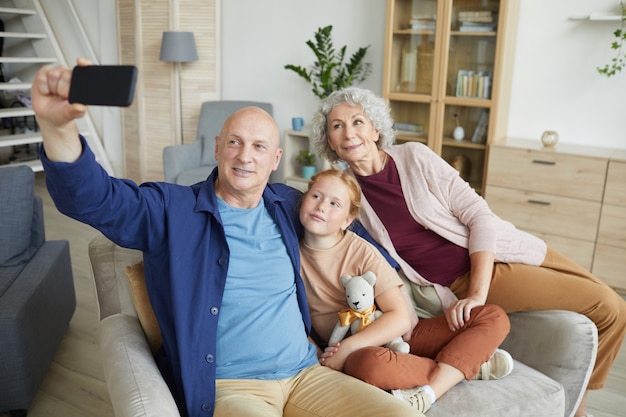 Portret van moderne senior paar selfie foto nemen via smartphone met schattige roodharige meisje in interieur