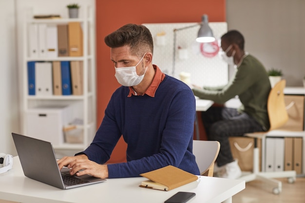 Portret van moderne volwassen man masker dragen en met behulp van laptop tijdens het werken aan de balie in kantoor, kopieer ruimte