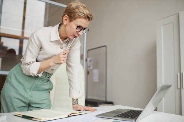 Portret van moderne zakenvrouw leaing over bureau terwijl kijken naar laptop scherm in kantoor, kopieer ruimte