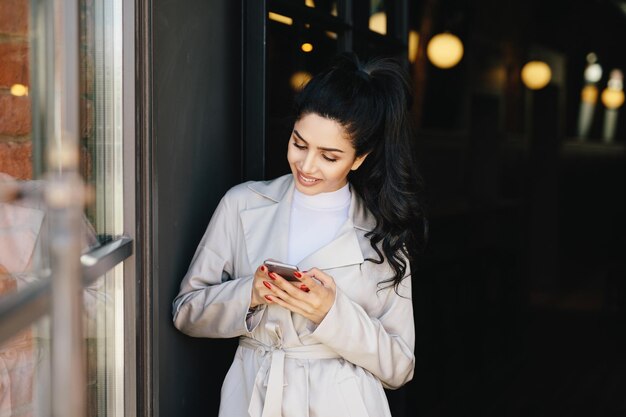 Portret van modieuze brunette vrouw met aangename uitstraling met mooie manicure met witte regenjas met smartphone in haar handen met aangename glimlach en blij om bericht te ontvangen