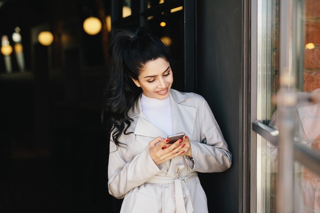 Portret van modieuze brunette vrouw met aangename uitstraling met mooie manicure met witte regenjas met smartphone in haar handen met aangename glimlach en blij om bericht te ontvangen