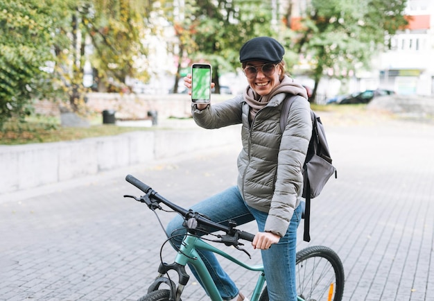 Portret van modieuze gelukkige jonge mooie vrouw in pet en zonnebril met behulp van mobiel op de fiets op zonnige herfstdag in stadspark mobiele mockup smartphone met groen scherm