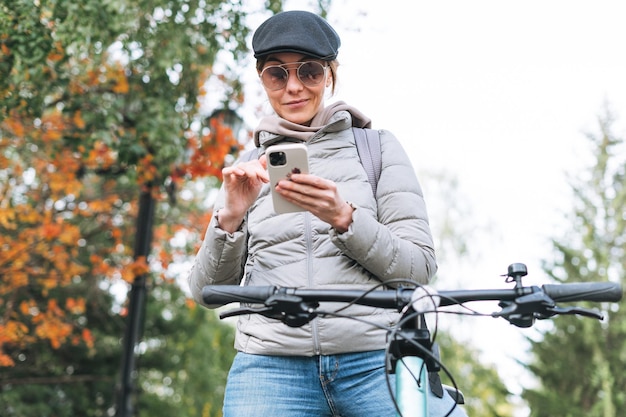 Portret van modieuze jonge mooie vrouw in pet en zonnebril met behulp van mobiel op de fiets op zonnige herfstdag in stadspark