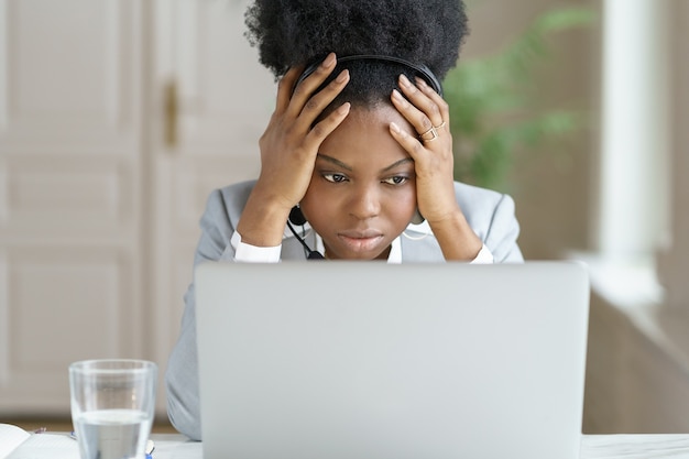 Portret van moe geïrriteerde afro zakenvrouw, koptelefoon dragen, praten met een slechte klant scherm laptop in kantoor kijken. Beambte verward door bericht van een ontevreden koper, hoofd aanraken