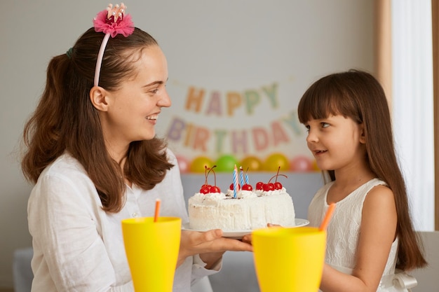 Portret van moeder en meisje die verjaardag vieren met heerlijke taart voor haar dochtertje, wachtend om de kaarsen te blazen, glimlachend, elkaar liefdevol en zachtaardig aankijkend.