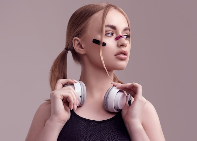 Foto portret van mooi blond meisje met glamourpleisters op haar gezicht luisteren muziek in witte hoofdtelefoons bij studio