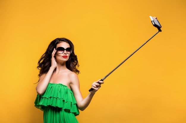 Portret van mooi brunette in groene kleding en rode lippen die zonnebril dragen terwijl het maken van selfie met stok