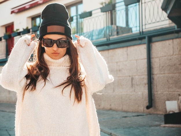 Foto portret van mooi lachend model. vrouw gekleed in warme hipster witte trui en muts. ze poseert op straat
