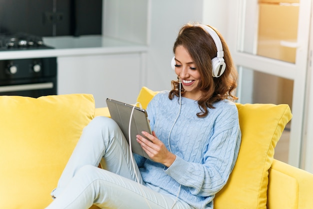 Portret van mooi meisje dat aan muziek met tablet op bank thuis luistert.
