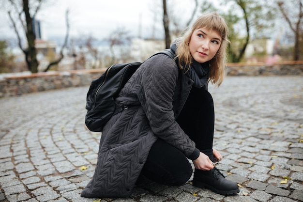 Portret van mooi meisje met blond haar dat bedachtzaam opzij kijkt terwijl gebogen op straat om de schoenveters te binden