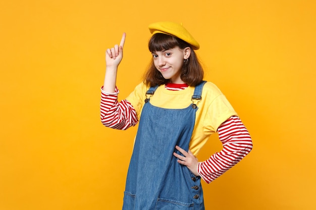 Portret van mooi meisje tiener in Franse baret, denim zomerjurk wijzende wijsvinger omhoog geïsoleerd op gele muur achtergrond in studio. Mensen oprechte emoties, lifestyle concept. Bespotten kopie ruimte.