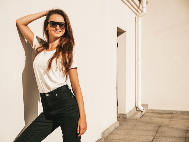 Portret van mooi model in zonnebril. vrouw gekleed in zomer hipster wit t-shirt en jeans. trendy vrouw poseren in de buurt van muur in de straat