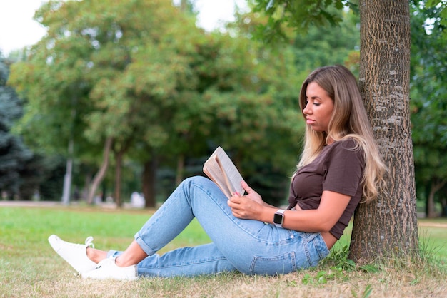 Portret van mooi mooi meisje lezer, jonge vrouw, student leest interessant boek in een zomerpark, zittend op de grond, groen gras, leunend op boom.