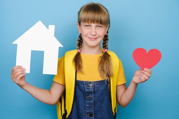 Portret van mooie aantrekkelijke lachende schoolmeisje met klein rood hart en papier wit huis model, gelukkig kijken naar de camera, draagt gele rugzak, geïsoleerd over blauwe kleur achtergrond in studio