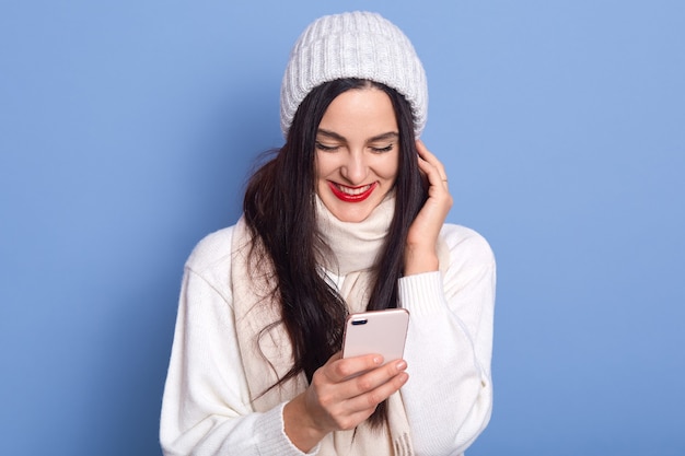 Portret van mooie aantrekkelijke tevreden vrolijk meisje in handen houden en het gebruik van mobiele telefoon, leest blij nieuws, dame staat geïsoleerd op blauwe ruimte