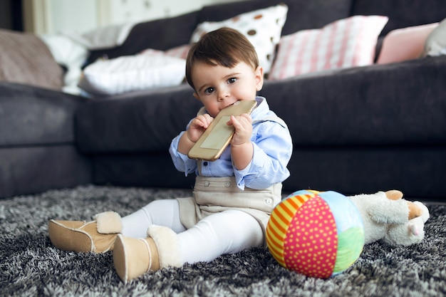 Portret van mooie baby die mobiele telefoon bijt terwijl hij thuis op de vloer van de woonkamer zit.
