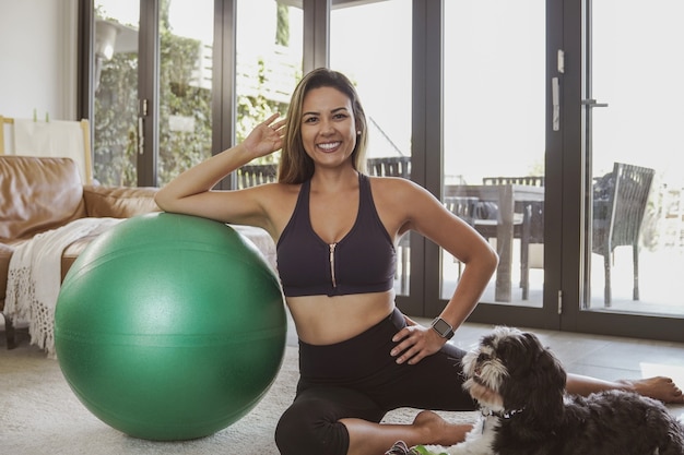 Foto portret van mooie braziliaanse vrouw in sportkleding met fitness bal en haar hond, thuis oefening training, online fitness klasse concept