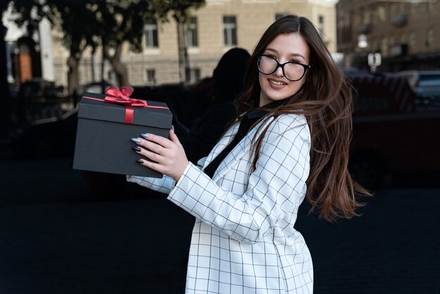 Portret van mooie brunette meisje met zwarte geschenkdoos in haar handen. Gelukkig stijlvol meisje houdt doos met rode strik.