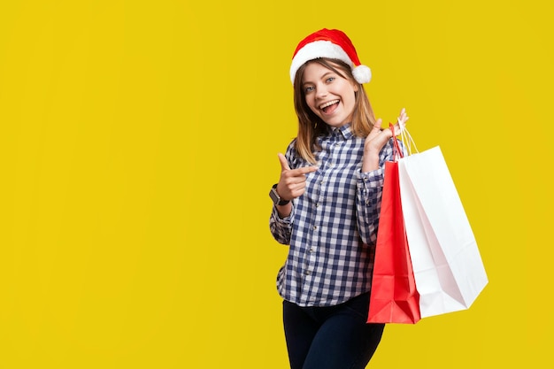 Portret van mooie brunette vrouw met nieuwjaar kerstmuts glimlach in casual shirt staande wijzend op boodschappentassen, tevreden kijkend met de aankoop. indoor studio-opname geïsoleerd op gele achtergrond