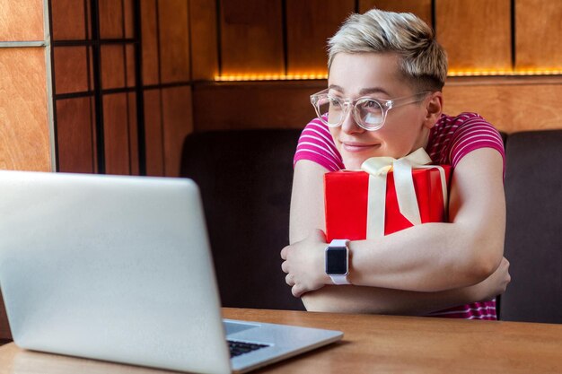 Portret van mooie dromende gelukkige jonge vrouw met kort haar in roze t-shirt en bril zit in café, knuffelt rode huidige doos, kijkt naar laptop en glimlacht. binnen