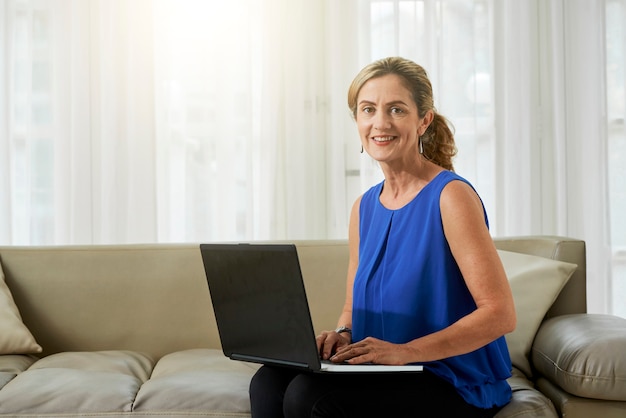 Portret van mooie elegante rijpe vrouw die thuis op de bank zit en op laptop werkt