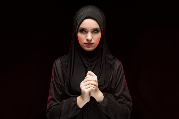 Portret van mooie ernstige jonge moslimvrouw die zwarte hijab met hand op hand dragen als het bidden concept op zwarte achtergrond