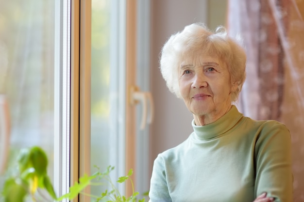 Portret van mooie glimlachende hogere vrouw met krullend wit haar. De bejaarde dame bevindt zich thuis door venster.