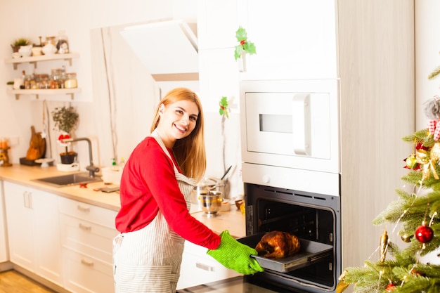 Portret van mooie glimlachende huisvrouw die thuis kookt. Moderne stijlvolle witte keuken, ideaal voor thuis koken en bakken. Kerst kalkoen, geroosterde kip