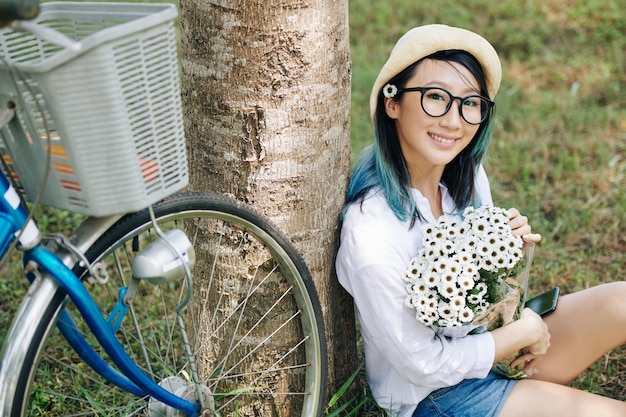 Portret van mooie glimlachende jonge Chinese vrouw die met boeket van madeliefjes onder boom zit