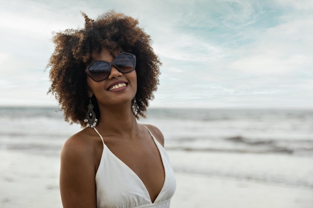 Portret van mooie glimlachende zwarte vrouw met krullend haar die een zonnebril op het strand draagt