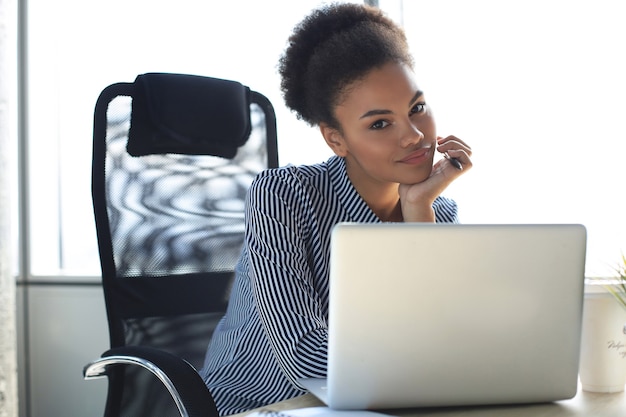 Portret van mooie jonge afro-amerikaanse vrouw die met laptop werkt terwijl ze aan tafel zit.