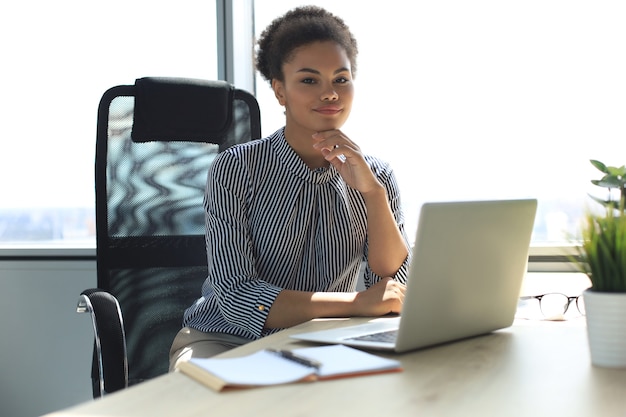 Portret van mooie jonge Afro-Amerikaanse vrouw die met laptop werkt terwijl ze aan tafel zit.