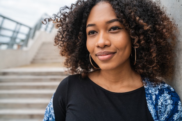 Foto portret van mooie jonge afro-amerikaanse vrouw met krullend haar staande tegen betonnen trap buitenshuis.