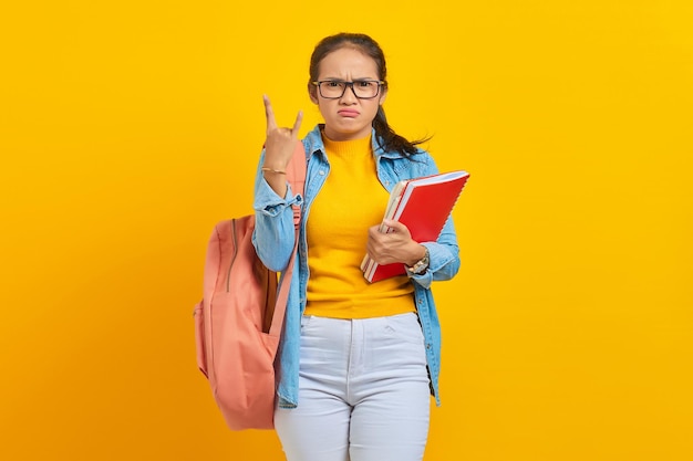 Portret van mooie jonge Aziatische student in denim kleding met rugzak met notitieboekje en schreeuwen met gekke uitdrukking doen rock symbool met handen omhoog geïsoleerd op gele achtergrond