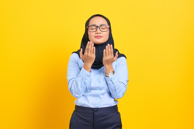 Portret van mooie jonge Aziatische vrouw die bidt met gesloten ogen, handpalmen naar boven gericht geïsoleerd op gele achtergrond