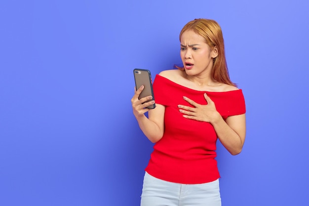 Portret van mooie jonge Aziatische vrouw in rode jurk met behulp van mobiele telefoon met verbaasde en geschokte uitdrukking geïsoleerd op paarse achtergrond