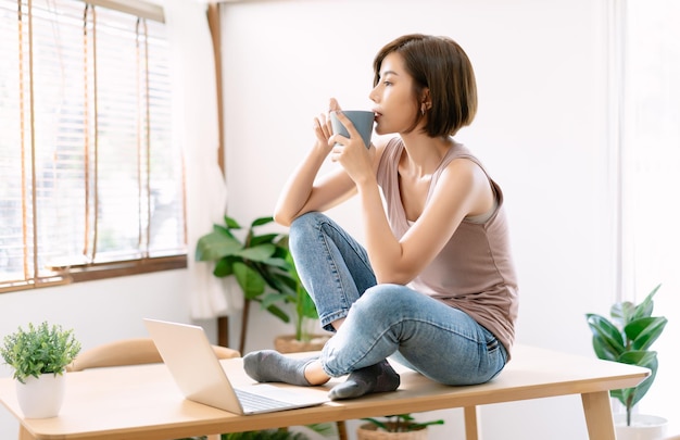 Portret van mooie jonge Aziatische vrouw ontspannen zittend op bureau genietend van haar kopje koffie en bedachtzaam uit het raam kijken Meisje tijdens werkpauze Genieten van tijd thuis