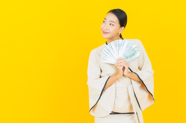 Portret van mooie jonge bedrijfs Aziatische vrouw met heel wat contant geld en spaarvarken op gele muur