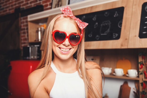 Portret van mooie jonge blonde lange haarvrouw die een hartenzonnebril draagt die op de camera kijkt en in de rode comfortabele zonnige keuken glimlacht