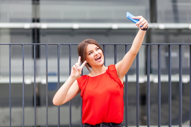 Portret van mooie jonge brunette vrouw in stijlvol rood shirt die selfie op smartphone neemt en overwinning of vredesgebaar toont, succes vierend. videogesprek voeren, bij het hek buiten staan