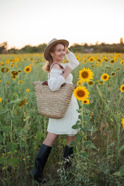 Portret van mooie jonge vrouw 33 jaar oud in hoed in zonnebloemveld bij zonsondergang Gelukkig model in witte jurk op zomeravond in de natuur Warm