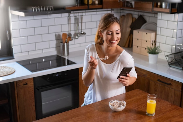 Portret van mooie jonge vrouw die ontbijt in de keuken en telefoon gebruikt voor sociale media