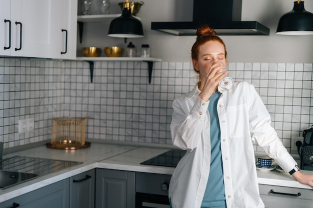 Portret van mooie jonge vrouw die witte wijn drinkt die in lichte keukenruimte staat