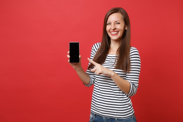 Portret van mooie jonge vrouw wijzende wijsvinger op mobiele telefoon met leeg zwart leeg scherm geïsoleerd op heldere rode achtergrond. Mensen oprechte emoties, lifestyle concept. Bespotten kopie ruimte.