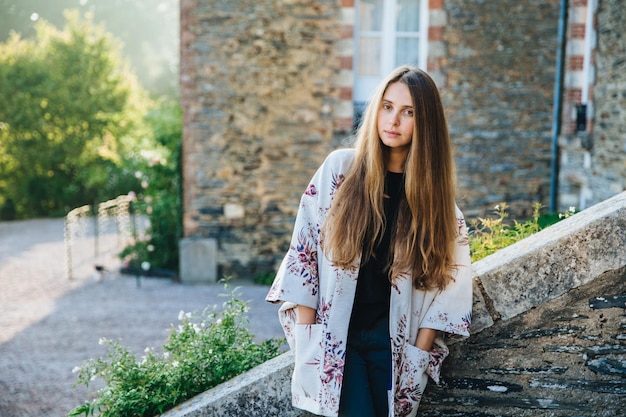 Portret van mooie jonge vrouwelijke model draagt warme mantel houdt handen in zak poses tegen oude monumenten geniet van prachtige natuur en rustige sfeer bewondert architecturale gebouwen