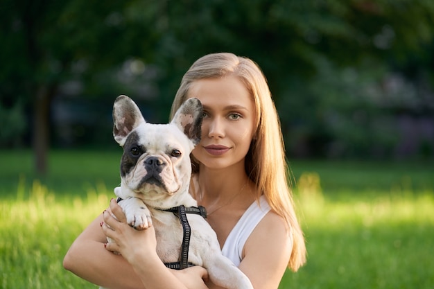 Portret van mooie kaukasische jonge vrouw die volwassen franse buldog houdt en direct in de zomerpark kijkt.