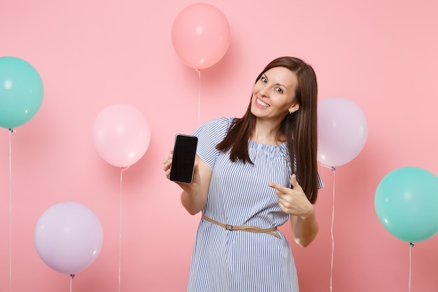 Portret van mooie lachende gelukkige vrouw in blauwe jurk met wijzende wijsvinger op mobiele telefoon met leeg leeg scherm op roze achtergrond met kleurrijke luchtballonnen. Verjaardag vakantie feest.