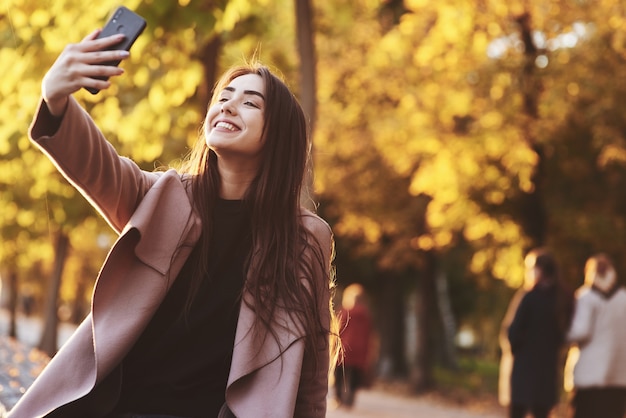 Portret van mooie, lachende jonge brunette meisje selfie maken met een zwarte smartphone, casual jas dragen in een zonnige, gele herfst tuin op de onscherpe achtergrond.